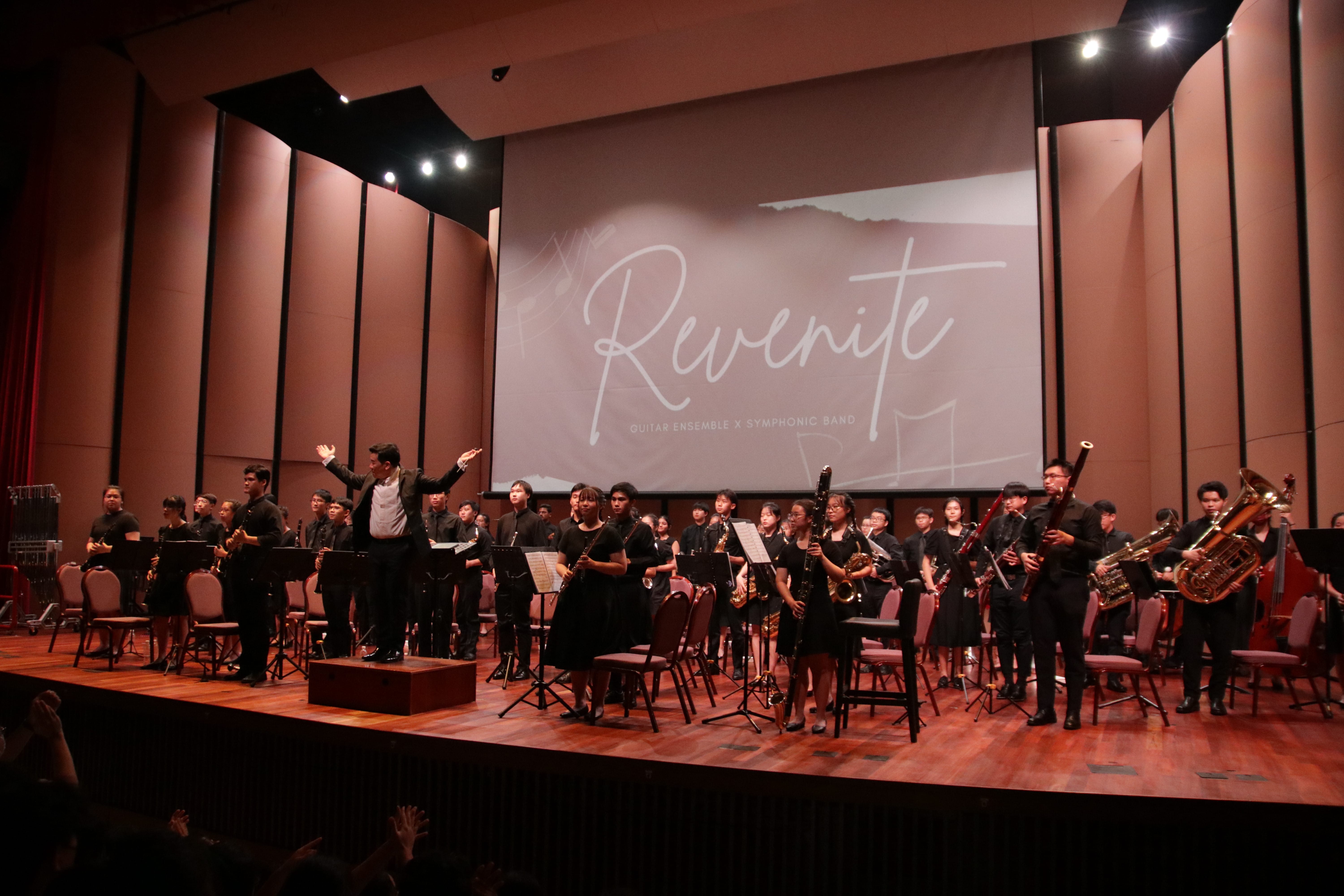 A conductor with raised arms leads a large youth symphonic band on stage, with a screen reading "Revenite" behind them.
